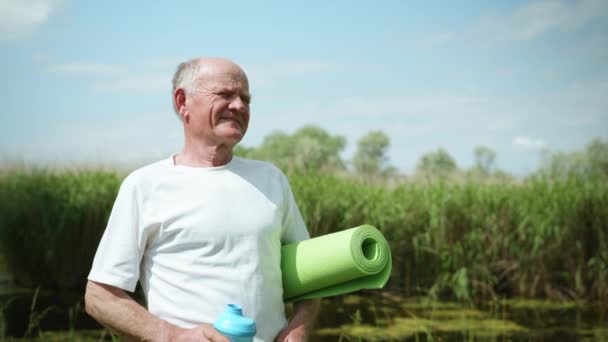 ヨガや瞑想のためのラグを持つ老人の肖像画を手に健康の世話をし、スポーツをした後、きれいな健康的な水を飲む - 映像、動画