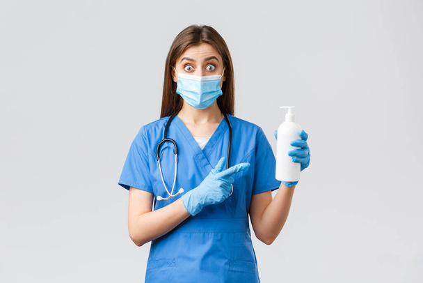 Covid-19, prevención de virus, salud, trabajadores sanitarios y concepto de cuarentena. Enfermera sorprendida, linda doctora en uniformes azules, máscara médica y guantes apuntando con el dedo a la mano desinfectante o jabón
 - Foto, Imagen