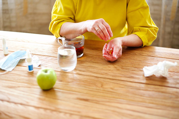 Side view of crop middle aged female taking pills while sitting at table with cup of tea and protective mask at home - Photo, Image