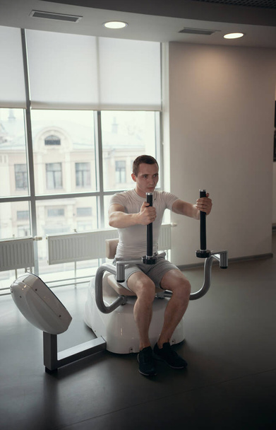 Modern gym - a guy sitting on the strength training machine and pumping his hands muscles - Zdjęcie, obraz
