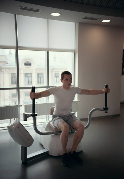 Academia moderna - um cara de camiseta branca sentado na máquina de treinamento de força e bombeando os músculos das mãos
 - Foto, Imagem
