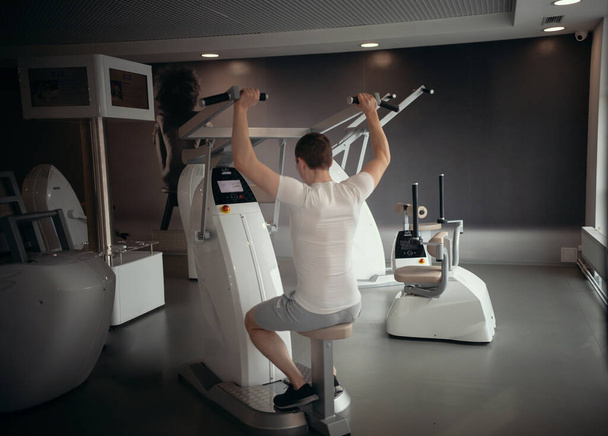 Modern jimnastik - beyaz tişörtlü genç bir adam. Güç antrenman makinesinin üzerinde oturuyor ve ellerini kasıyor. - Fotoğraf, Görsel
