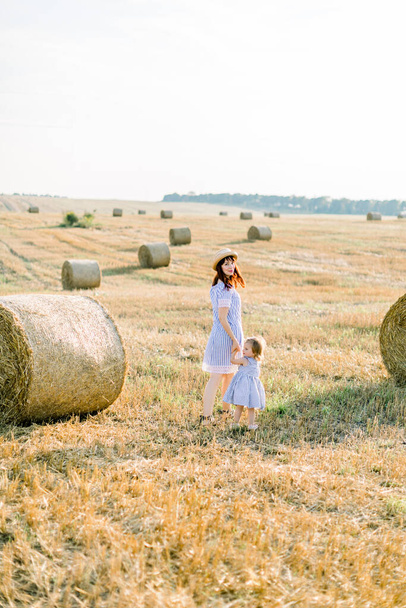 Summer bright portrait of little baby girl playing with her mom, wearing similar striped dresses, in beautiful field with hay bales on the background. summer, sunny walking, happy childhood - Foto, Imagem
