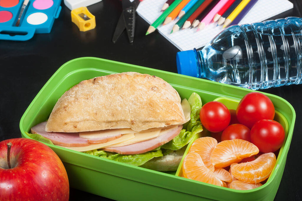 School lunch. Sandwich, small tomatoes, tangerine, apple in plastic lunch box and bottle of water on black chalkboard. - Photo, Image