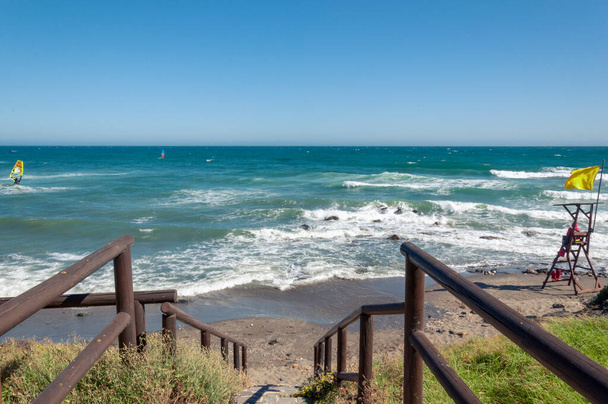 Un escalier en pierre avec rampe en bois mène à une plage rocheuse, un sauveteur est assis sur une tour avec un drapeau jaune et regarde le kitesurfer, la mer avec des vagues et le vent
. - Photo, image