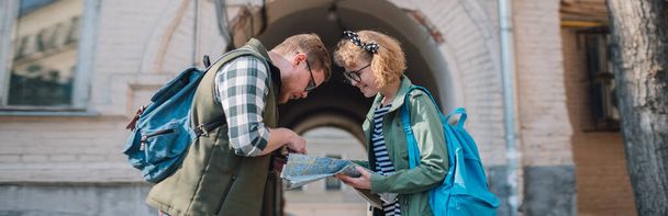 Пара туристов в европейском городе с картой. Молодой человек и женщина с туристической картой в руках идут по европейским улицам. Поиск маршрута путешествия на бумажной карте
 - Фото, изображение