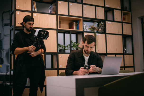 Filming a blog, workshop or live broadcast. A man with a camera shoots a guy at a laptop in a workspace. Professional cameraman and speaker make video in coworking - Fotografie, Obrázek