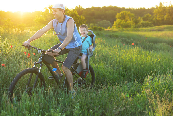 Happy father and child in bicycle chair riding by the meadow in beautiful landscape. Healthy Lifestyle Concept. - 写真・画像