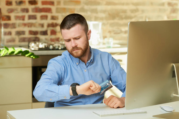 Un hombre con barba frunce el ceño y sostiene una tarjeta de crédito mirando su reloj de pulsera en casa. Un hombre que espera tiempo para hacer un pago en línea en Internet
. - Foto, Imagen