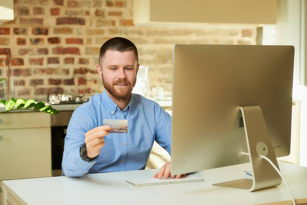 Мужчина с бородой набирает номер кредитной карты для покупок дома. Парень делает онлайн-платеж в интернете на настольном компьютере в своей квартире
. - Фото, изображение