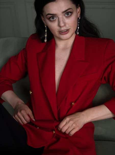 Красивая юная брюнетка в красной куртке сидит на сером кресле на сером фоне
 - Фото, изображение