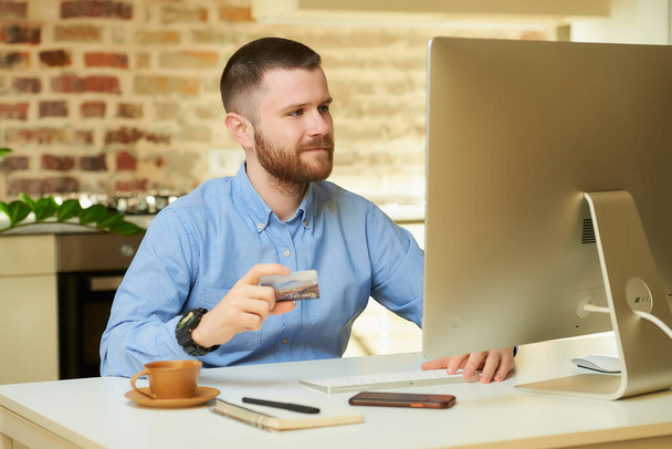 髭を生やした男がコンピュータの前に座り、自宅でクレジットカードを持っている。彼のアパートのデスクトップコンピュータ上のインターネット上でオンラインで支払いをしている男.  - 写真・画像