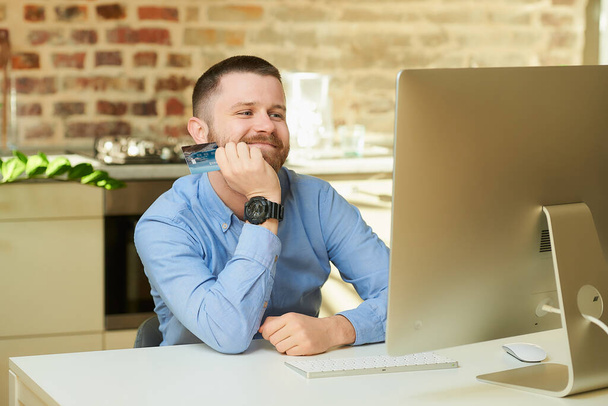 Un hombre feliz con barba espera delante de la computadora y tiene una tarjeta de crédito en casa. Un tipo haciendo un pago en línea en Internet en una computadora de escritorio en su apartamento
. - Foto, imagen
