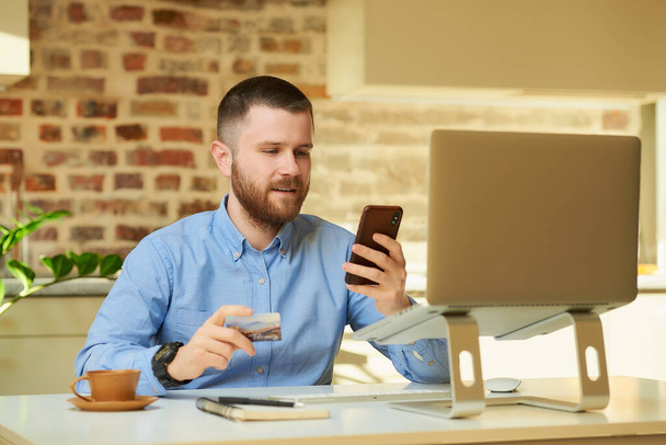 Мужчина ищет онлайн-продажи на смартфоне, держа кредитку в руке перед компьютером дома. Парень делает онлайн-платеж через интернет на ноутбуке в своей квартире
. - Фото, изображение