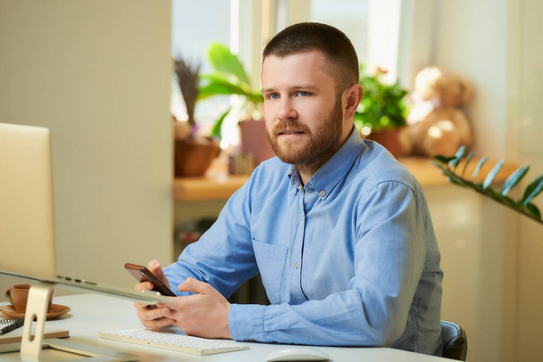 Босс слушает отчет о своих сотрудниках на онлайн-брифинге перед ноутбуком в его квартире. Мужчина в синей рубашке с бородой, удаленно работающий из дома
.  - Фото, изображение