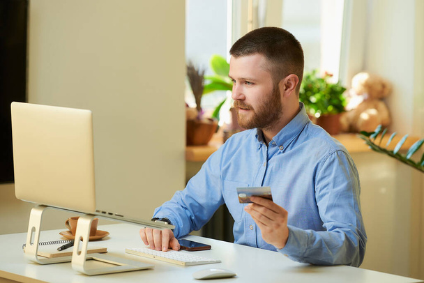 Ein Mann auf der Suche nach Produkten, die er in einem Online-Shop auf seinem Laptop mit seiner Kreditkarte zu Hause kaufen kann. Ein Mann mit Bart bei einer Online-Zahlung im Internet auf einem Laptop in seiner Wohnung. - Foto, Bild