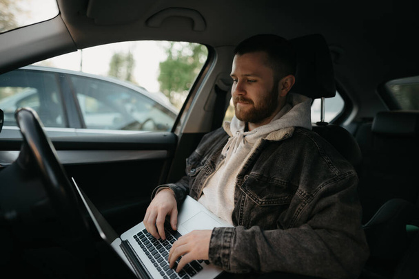 Um homem feliz com uma barba fazendo negócios on-line em seu computador portátil dentro de um carro de conforto. Um cara parou seu carro para resolver imediatamente remotamente tarefas no trabalho à distância social
.  - Foto, Imagem