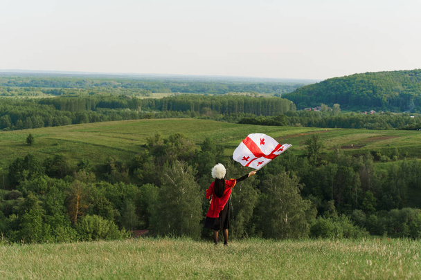 Грузинська дівчинка з національним прапором Грузії в руках ходить навколо зелених пагорбів. Грузинський спосіб життя. Жінка в Папахе і червоне плаття. - Фото, зображення