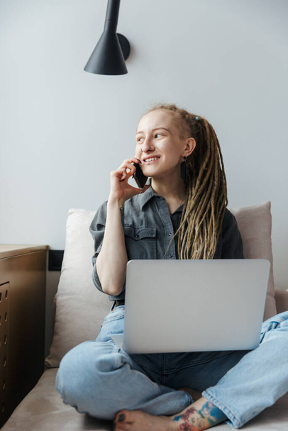 Image of a smiling young girl with dreadlocks and piercing indoors using laptop computer talking by mobile phone. - Φωτογραφία, εικόνα