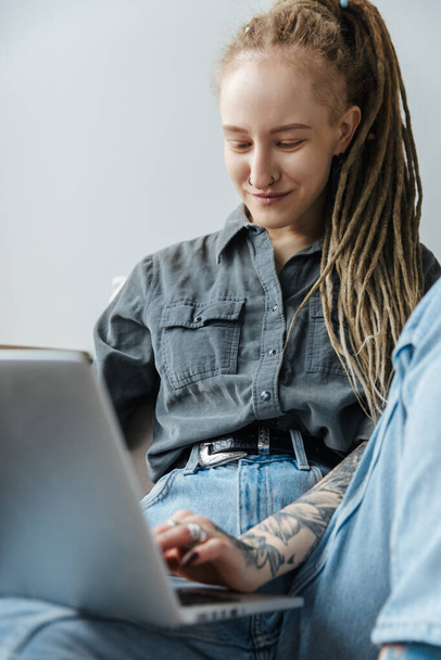 Image of a positive optimistic young girl with dreadlocks and piercing indoors using laptop computer. - Фото, изображение