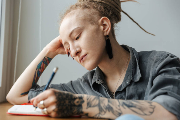 Image of a serious beautiful young girl with dreadlocks an piercing indoors writing notes in notebook. - Photo, image