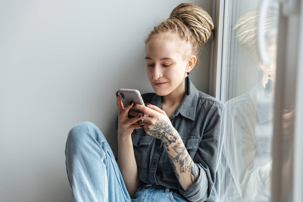 Image of a positive pleased young girl with dreadlocks and piercing indoors using mobile phone near window. - Foto, immagini