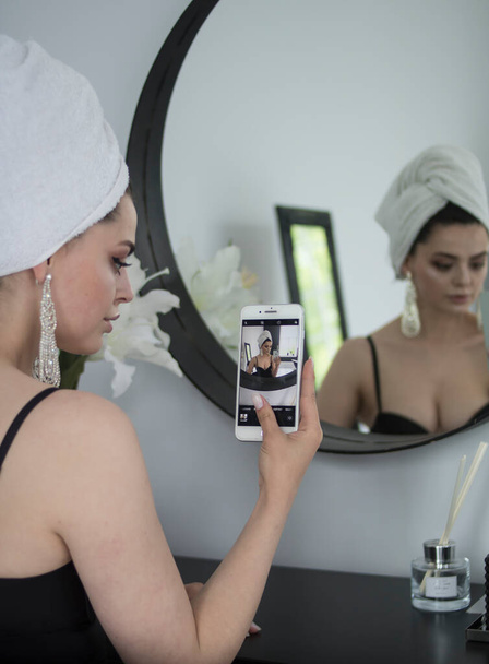 A young cute brunette girl with a white towel on her head and dressed in a black bodysuit with a phone in her hands sits near a mirror and takes a selfie - Zdjęcie, obraz