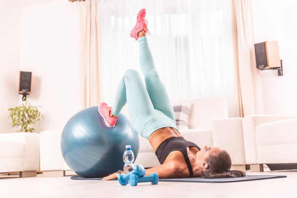 Female working out at home, lifting her core from the ground, one leg stretched, one leg on a fit ball. - Foto, imagen