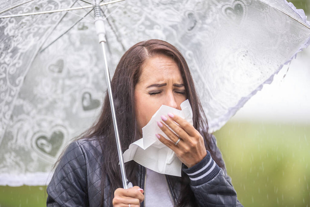 Woman with an umbrella outdoors sneezes into a napkin, getting cold from the bad weather. - Foto, Imagem
