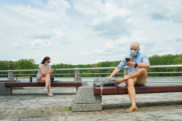 Een kale man en een vrouw in medische gezichtsmaskers met behulp van smartphones houden sociale afstand zittend op verschillende banken op een promenade. Een jongen en een meisje rusten in de sociale afstand in het park. - Foto, afbeelding