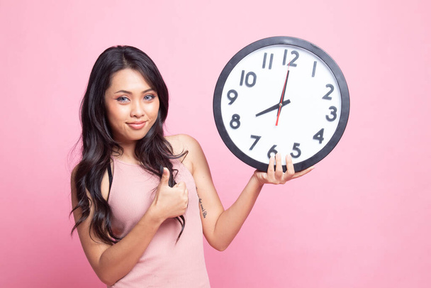 Young Asian woman thumbs up with a clock on pink background - Valokuva, kuva