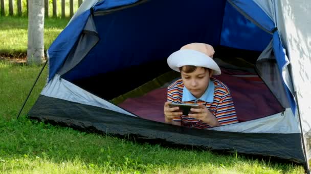 Şapkalı küçük çocuk çadırda oturmuş telefonuyla oyun oynuyor. Çocuk akıllı telefondan bir video izliyor. Taşınabilir İnternet. - Video, Çekim