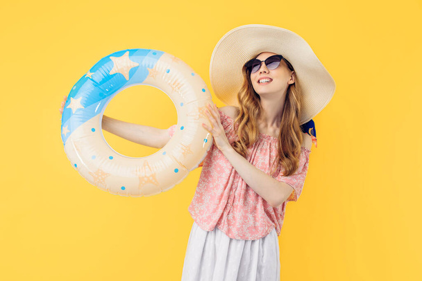 An attractive beautiful young woman in a straw hat standing in a floating inflatable ring and smiling isolated on a yellow background - Фото, зображення