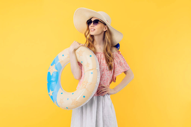 An attractive beautiful young woman in a straw hat standing in a floating inflatable ring and smiling isolated on a yellow background - Foto, Imagem