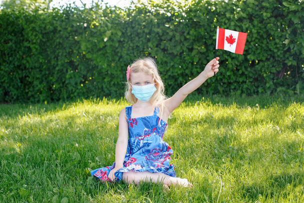 Menina caucasiana no rosto máscara protetora segurando acenando bandeira canadense ao ar livre. Criança criança em máscara sanitária na grama no parque celebrando o feriado do Dia do Canadá durante a epidemia de coronavírus.  - Foto, Imagem