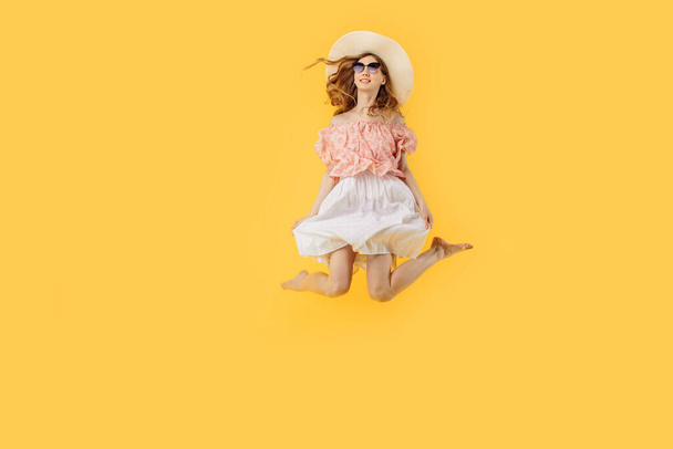 Full-length photo of a happy girl in a summer hat and sunglasses in summer, jumping high on an isolated yellow background - Фото, изображение