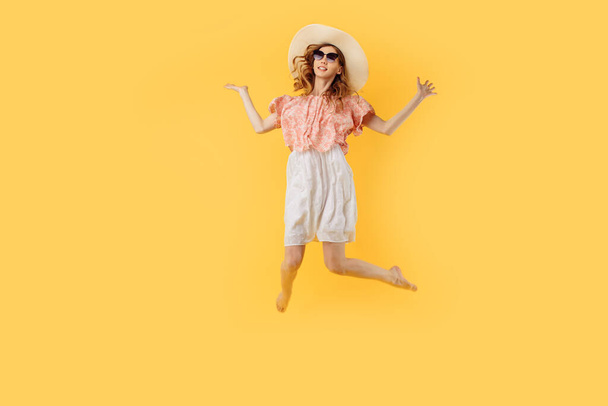 Full-length photo of a happy girl in a summer hat and sunglasses in summer, jumping high on an isolated yellow background - Photo, image
