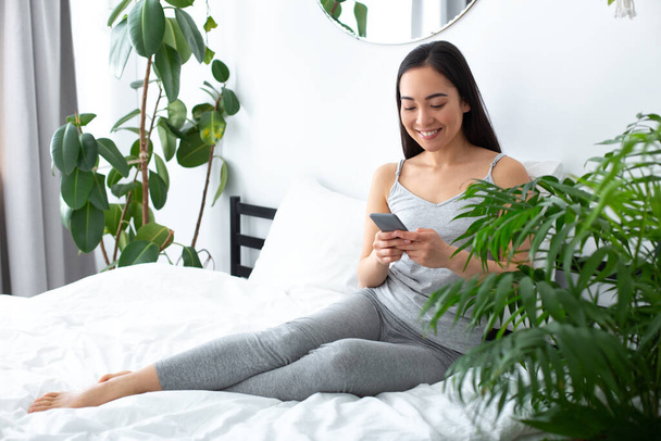 Happy attractive woman messaging on cell phone in bedroom - Photo, image