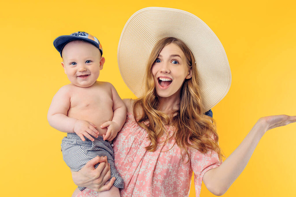 Happy shocked young mother in a summer hat holding a little boy on a yellow background. Summer holidays - Фото, изображение