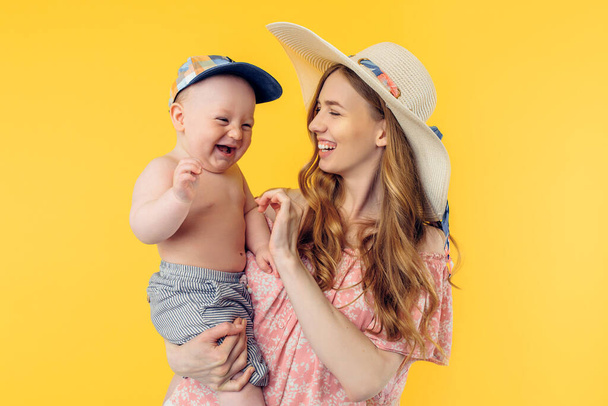 Happy young beautiful mother in a summer hat holding a little boy on a yellow background. Summer holidays - Foto, Imagem