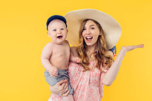 Happy shocked young mother in a summer hat holding a little boy on a yellow background. Summer holidays - Фото, зображення
