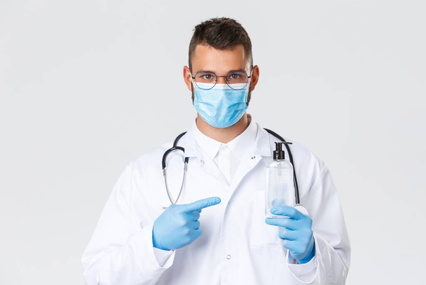 Covid-19, healthcare workers, pandemic and virus concept. Close-up of serious hispanic doctor in white scrubs and medical mask, gloves pointing finger at hand sanitizer, ask patients use it - Fotoğraf, Görsel