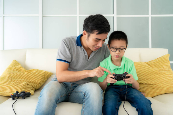 Батько азіатської сім'ї навчає сина грати в відеоігри з джойстиками, сидячи на дивані у вітальні, концепції сімейних цінностей та сучасної гри.. - Фото, зображення