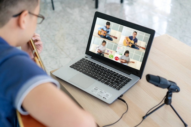 アジアの少年は、社会的距離のためのビデオ通話でオンライン会議のためのラップトップとビデオ会議で友人とオンラインで音楽を再生するためのアコースティックギターの仮想幸せな時間会議を演奏. - 写真・画像
