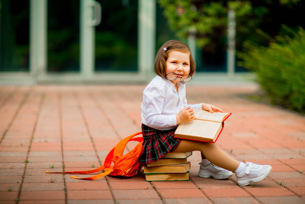 a little girl in a school uniform and glasses,sitting on a stack of books near the school - Фото, изображение