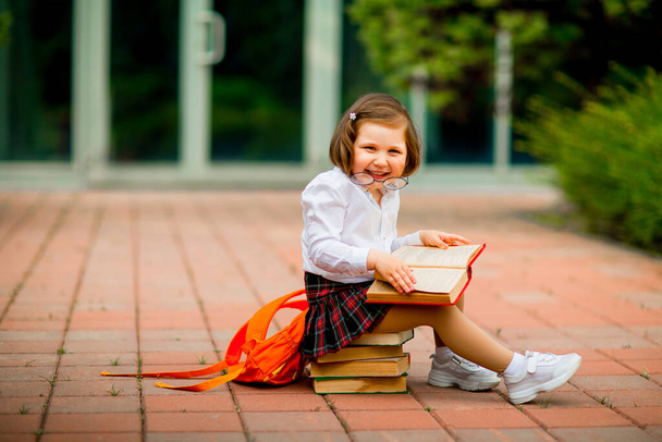 a little girl in a school uniform and glasses,sitting on a stack of books near the school - Fotografie, Obrázek