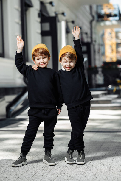 Two cheerful embraced twin boys in casual outfit posing with hands up in the street. - Zdjęcie, obraz