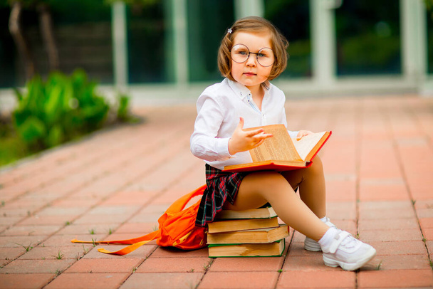 a little girl in a school uniform and glasses,sitting on a stack of books near the school - Фото, зображення