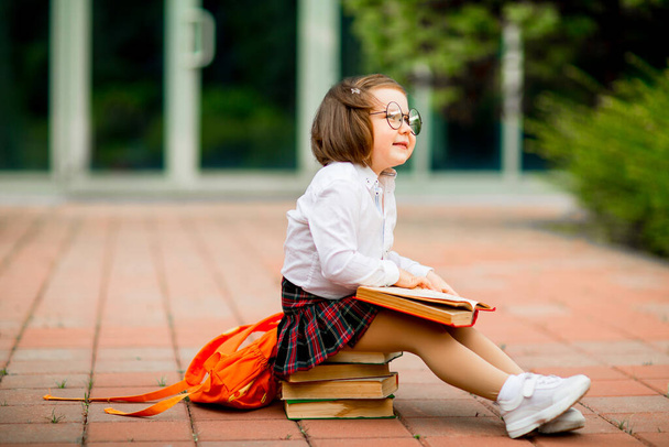 a little girl in a school uniform and glasses,sitting on a stack of books near the school - Foto, imagen