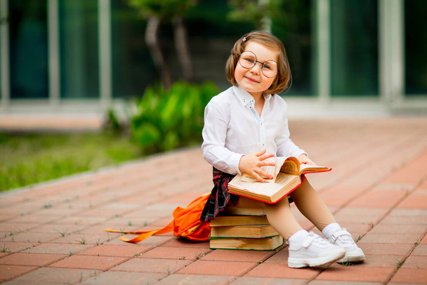 a little girl in a school uniform and glasses,sitting on a stack of books near the school - Foto, Imagem